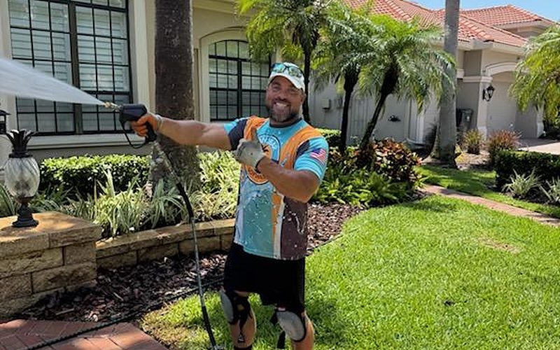 Smiling Sandbar crew member in a branded jersey rinsing a paver walkway at a tile-roof home — real job photo